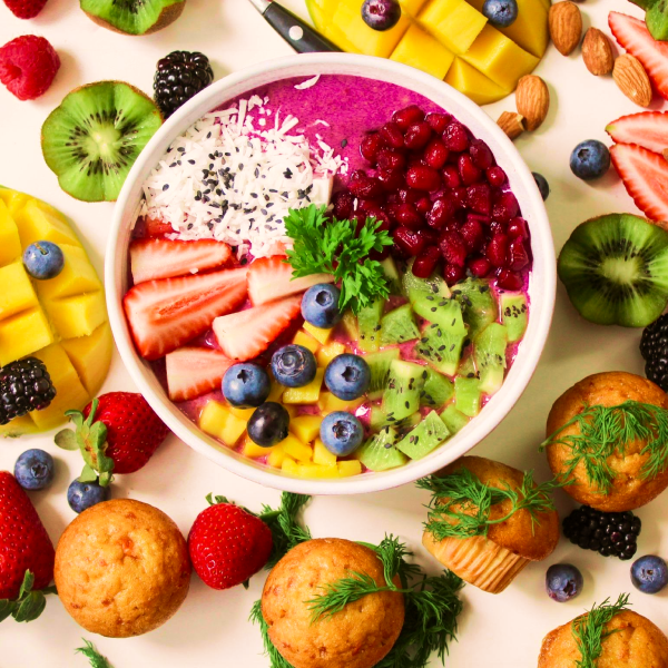 A white bowl filled with a brightly colored fruit salad, surrounded by fruits of various hues.