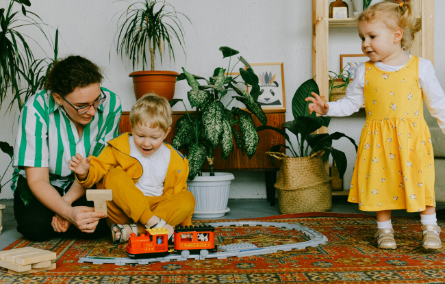 Two children dressed in yellow are playing with a toy train while their mother looks on happily.