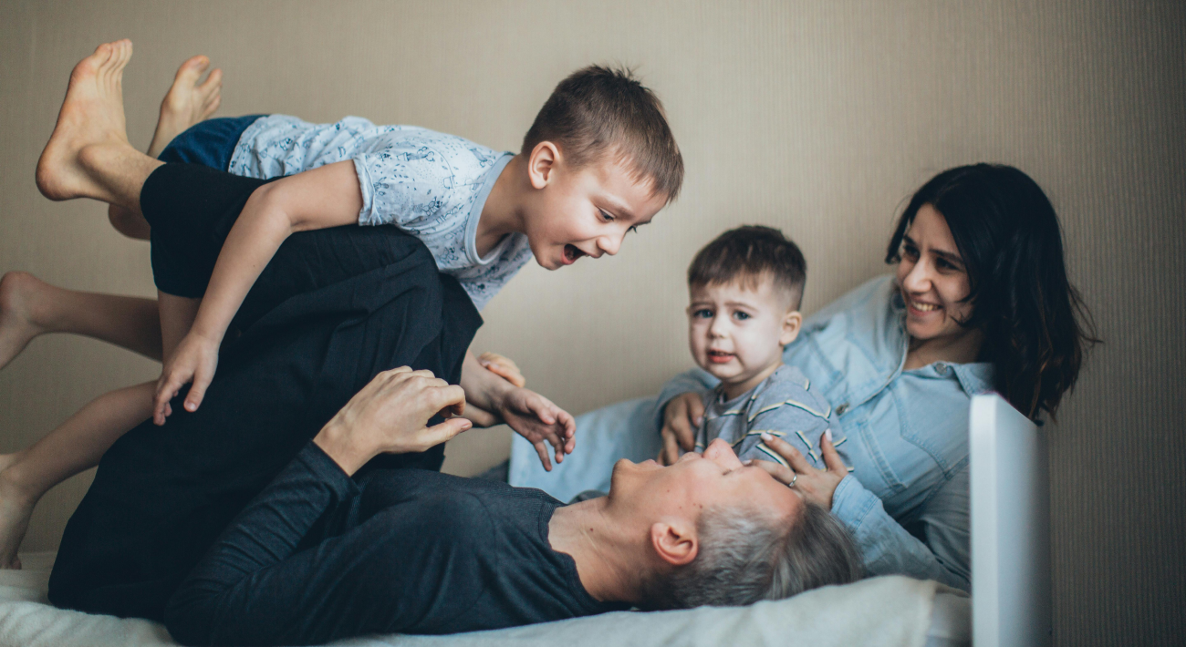 Father and son are playing on the bed while the mother and the little brother watch happily.