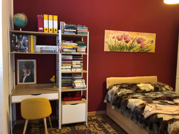 Feng Shui design by Raffaela Marescalco. A small bedroom with a burgundy wall. On the left side, there is a modest desk with an attached bookshelf and a yellow chair. On the right side is the bed, with a picture above it.
