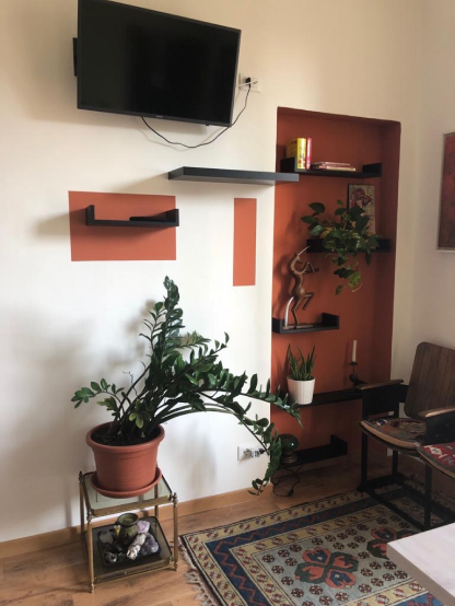 Feng Shui design. The room has white walls with burgundy accents. On the right, there are two small wooden chairs. A rug in soft colors covers the floor, and a potted plant in a terracotta pot is centered in the room.