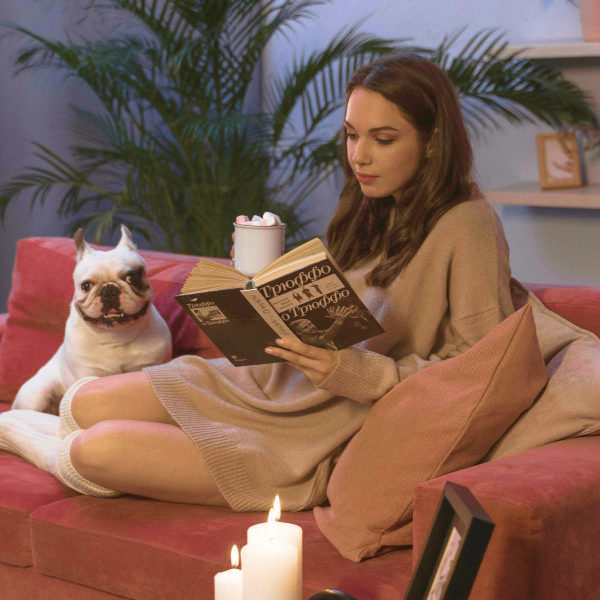 A girl is sitting on the sofa, reading a book while sipping hot chocolate. Her dog is also sitting on the sofa, watching her.