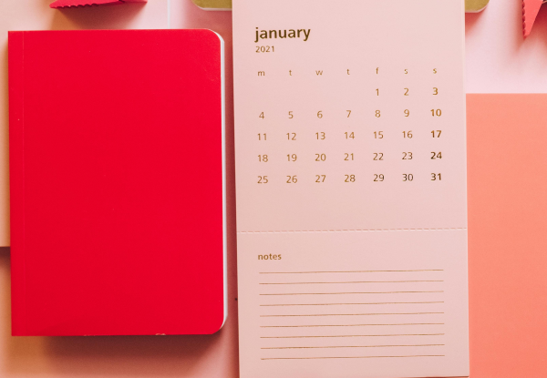 A calendar open on a light wooden table, next to a red notebook with a black pen on top. The calendar is monthly and shows January 2021. The calendar squares are empty except for a few dates circled in red. The notebook is closed with a smooth, even cover. The pen is open, and its cap lies next to it.