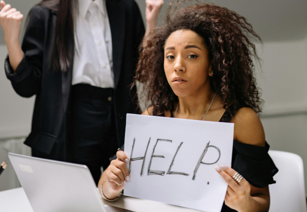 A girl holds a sheet with the word "HELP" after being bullied by the colleague to her right. The image represents karmic wounds and rebirth.