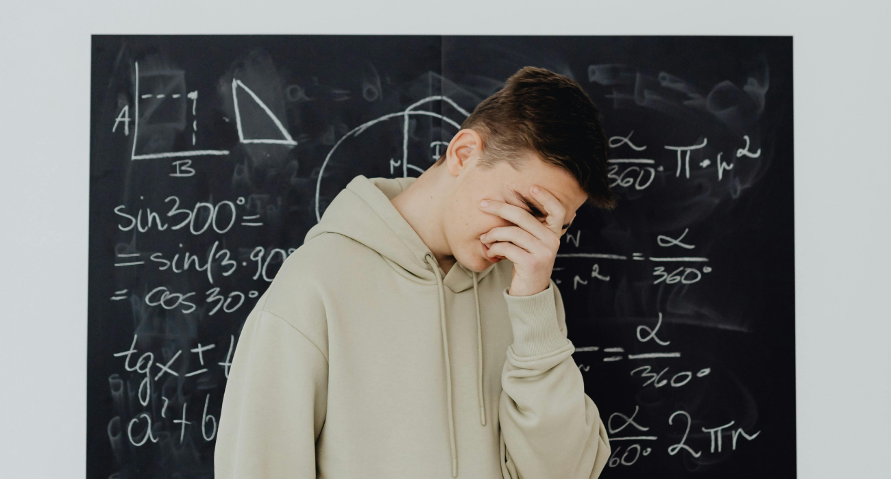 A boy in a sweatshirt, standing in front of a blackboard, looks disappointed with his academic performance and covers his face with his hand. Karmic wounds and rebirth.