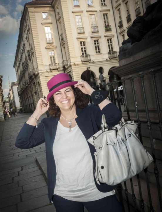 Raffaela Marescalco is sporting a smile as she poses in the center of Turin. She is dressed in a blue jacket and trousers, a white T-shirt, and a fuchsia hat, while carrying her pearl-colored bag. This is one of the outfits she wears for her Feng Shui consultations.