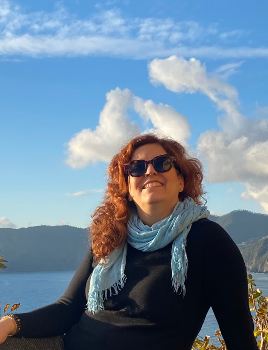 Raffaela Marescalco, smiling and wearing dark brown sunglasses, poses in front of a lake on a beautiful sunny day.