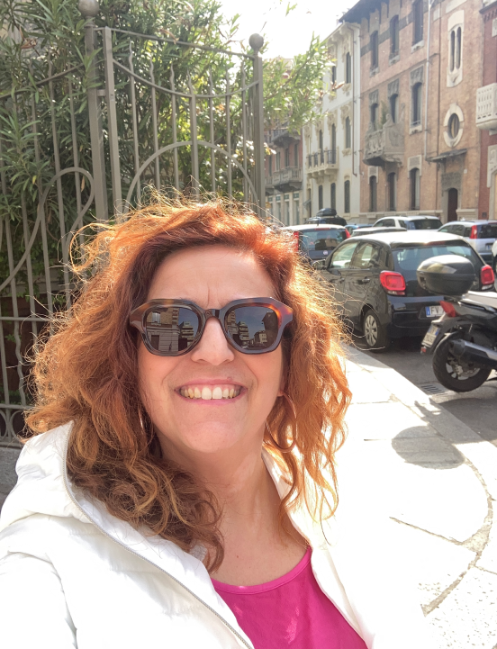 Raffaela Marescalco, smiling and wearing dark brown sunglasses, poses in front of a gate behind which there is a plant.