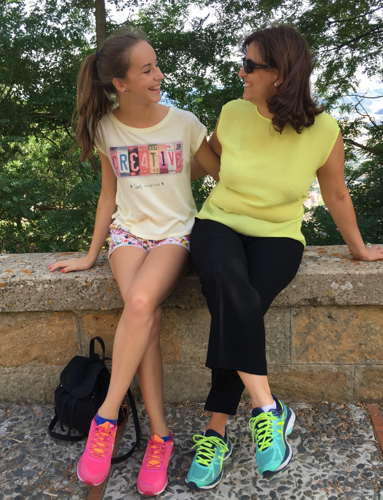Raffaela Marescalco and her older daughter Sarah are sitting on an outdoor wall, smiling as they look into each other's eyes. They are both dressed casually and wearing colorful sneakers.