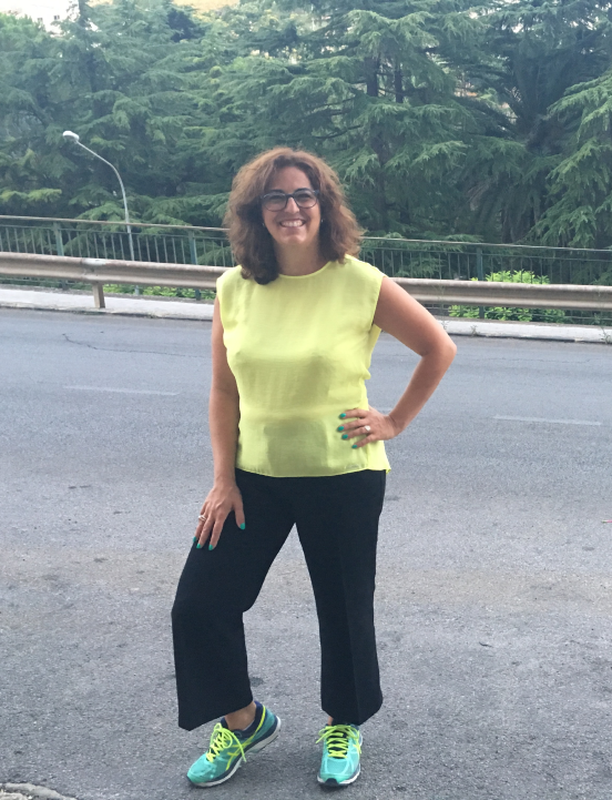 Raffaela Marescalco is posing in a running outfit. She is wearing a sleeveless yellow shirt, black long pants, and aqua green running shoes.