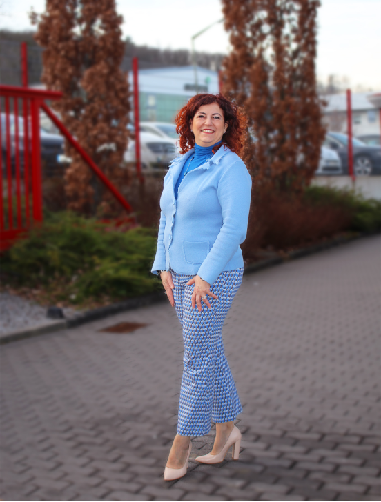 Raffaela Marescalco stands on a tree-lined avenue, wearing pants, a shirt, and a jacket in different shades of blue, paired with cream-colored heels.